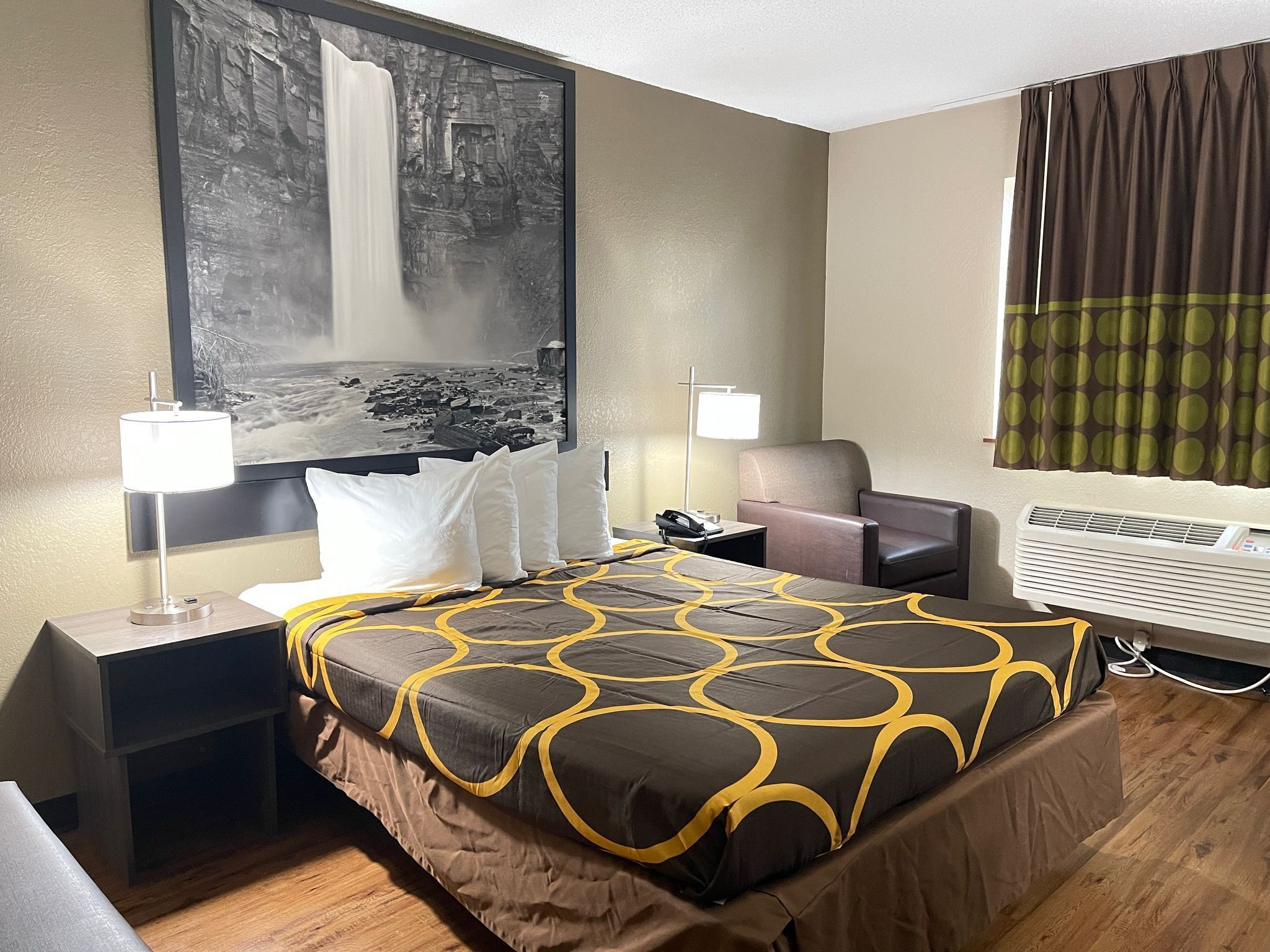 Habitación de hotel con cama, dos mesitas de noche, un escritorio, una silla, un cuadro grande, una ventana con cortinas y un equipo de aire acondicionado.
