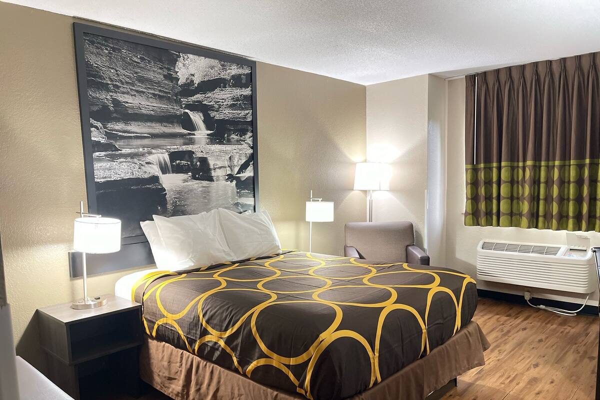 A hotel room with a bed, two bedside tables, a wall-mounted air conditioner, and a large black and white photograph.