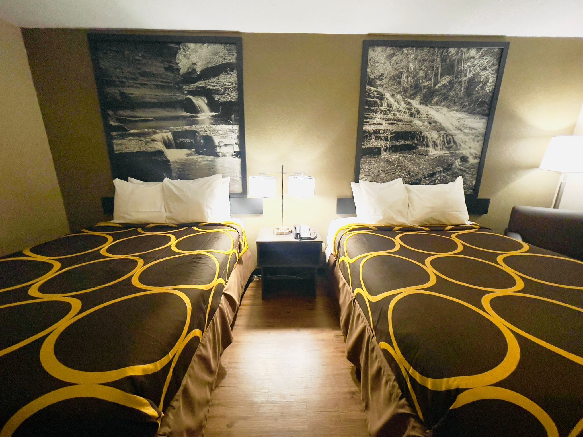 A hotel room with two beds, a nightstand, a lamp, and two large black and white photographs on the walls.