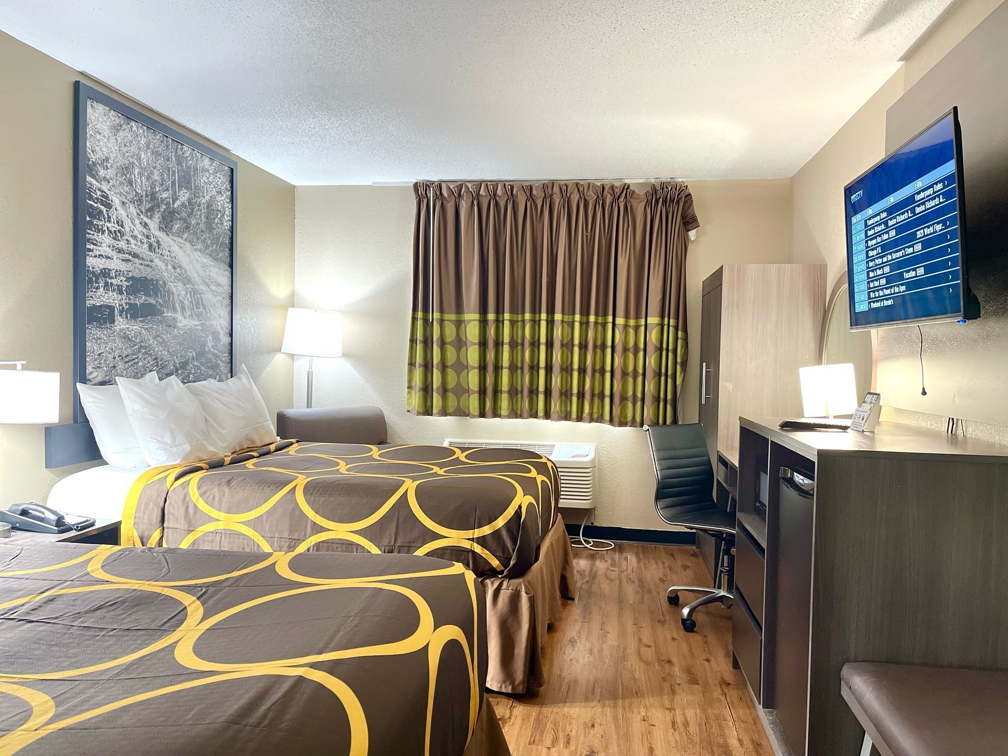 A hotel room with a bed, a desk, a chair, a TV, and a large painting on the wall.