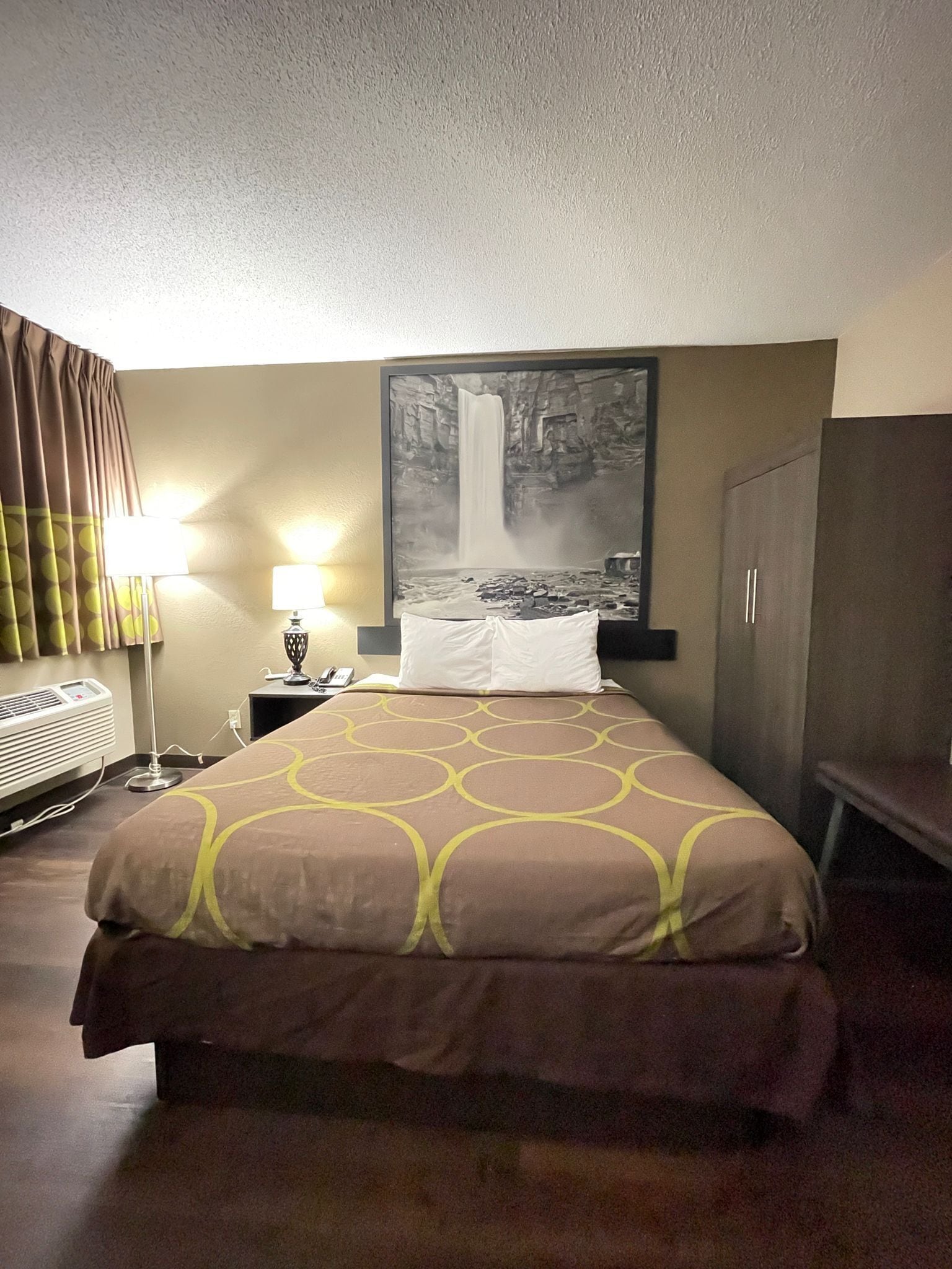 Camera d'albergo con un letto, un comodino con una lampada, un quadro che raffigura una cascata e un armadio.