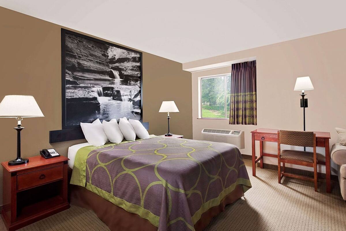 A hotel room with a bed, two bedside lamps, a desk with a chair, a large window, and a wall-mounted air conditioner.