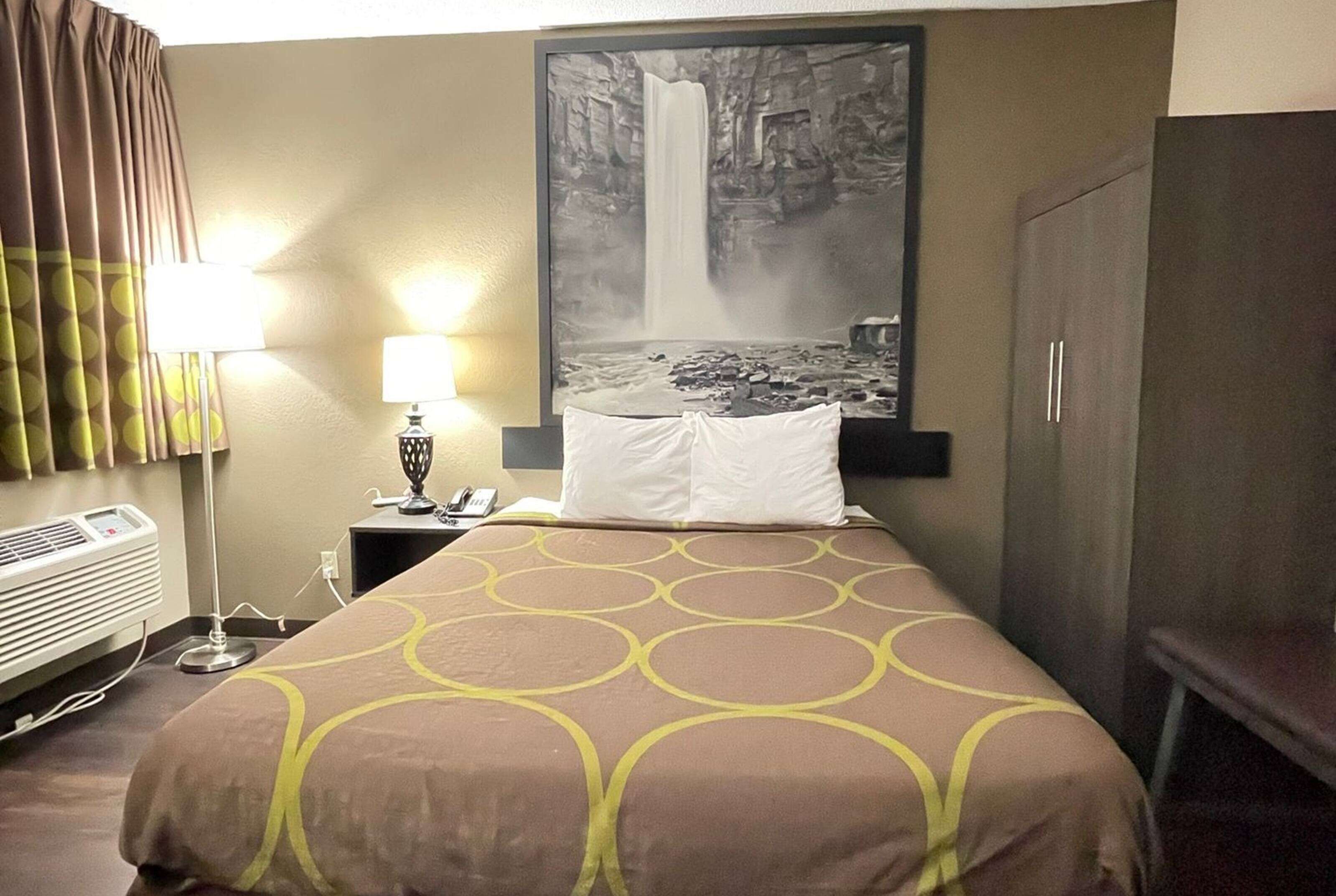 Ein Hotelzimmer mit einem Bett, einem Nachttisch mit Lampe, einem Wandbild von einem Wasserfall und einem Kleiderschrank.