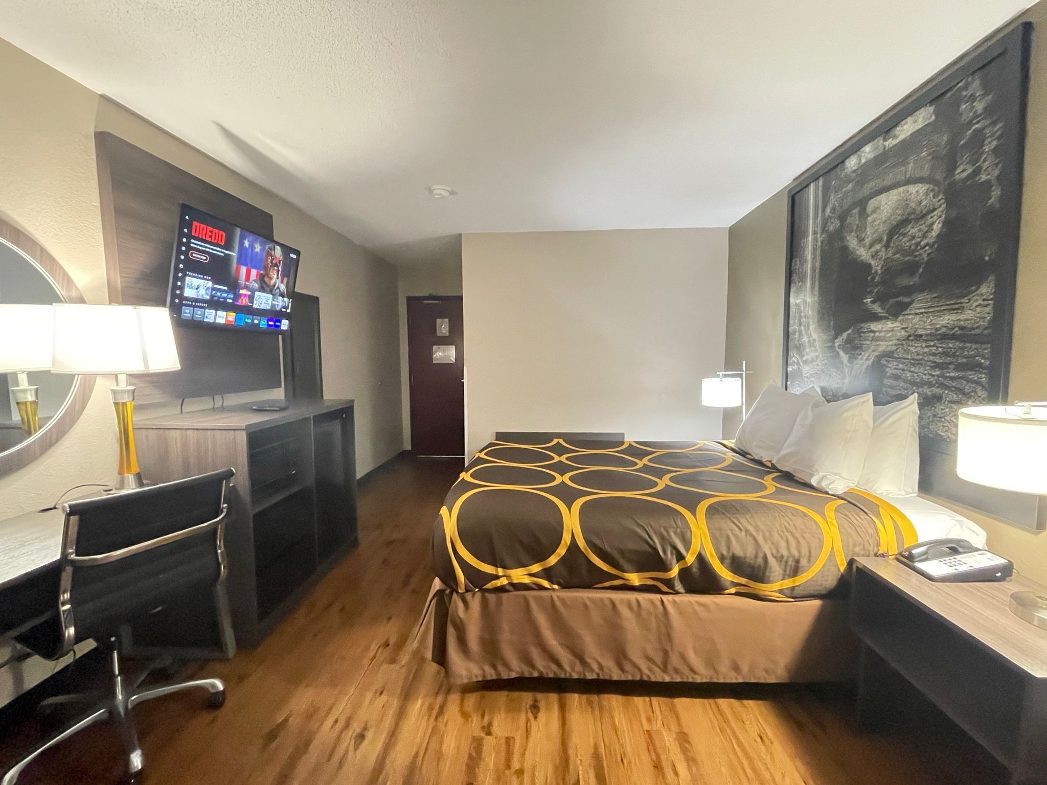 A hotel room with a bed, desk, chair, TV, and artwork on the wall.