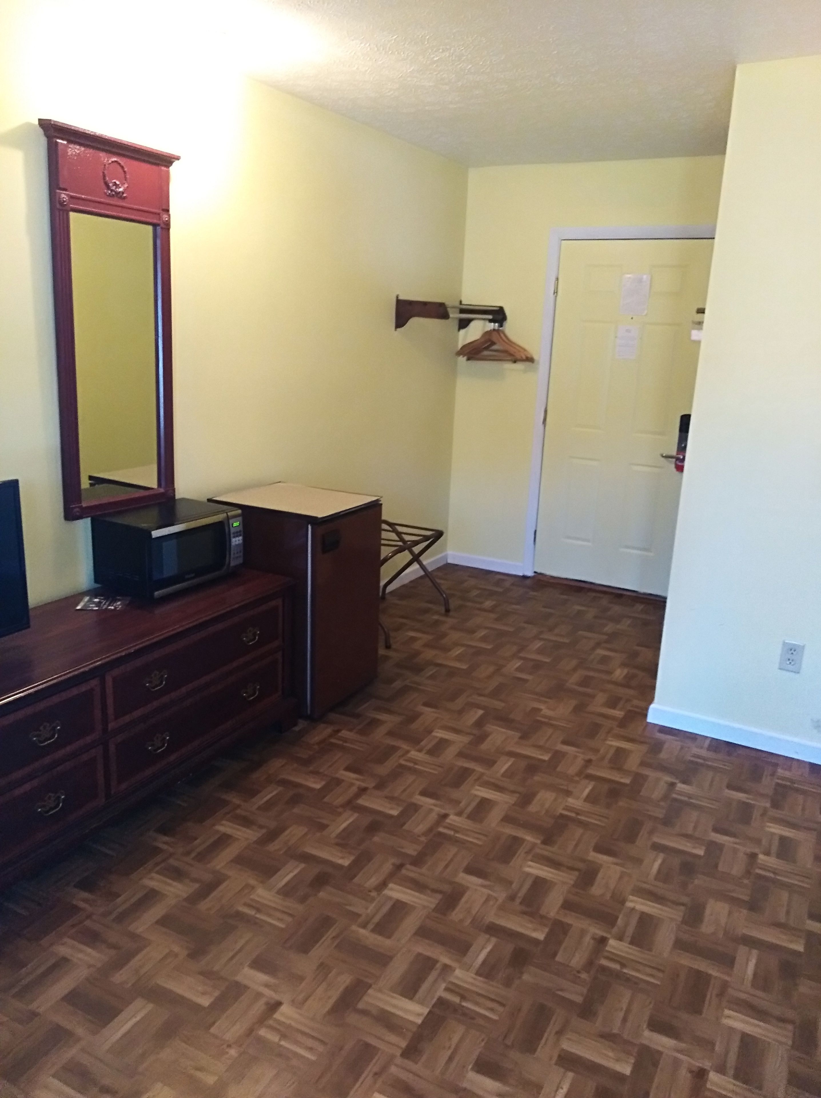 A room with a wooden herringbone floor, a dresser with a mirror, a microwave, and a coat rack with hangers.