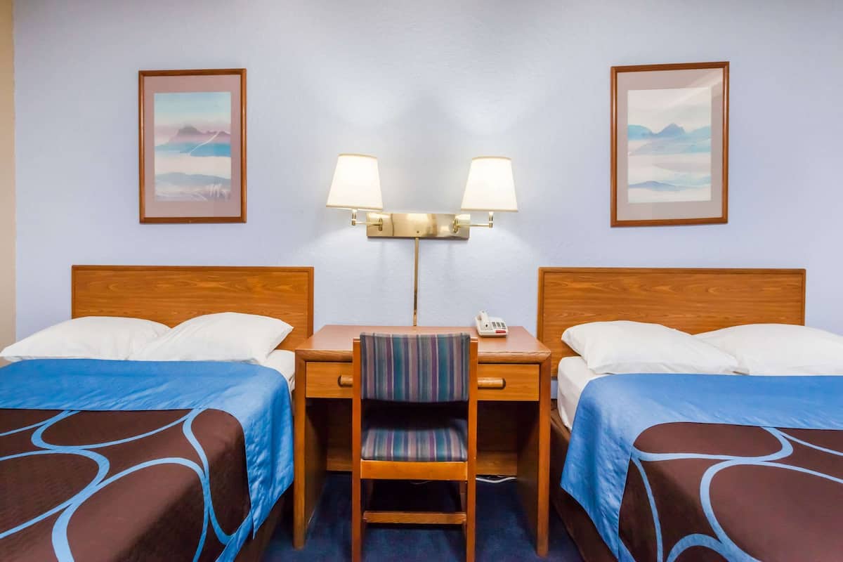 A hotel room with two beds, a desk with a chair, a lamp, and two framed pictures on the wall.