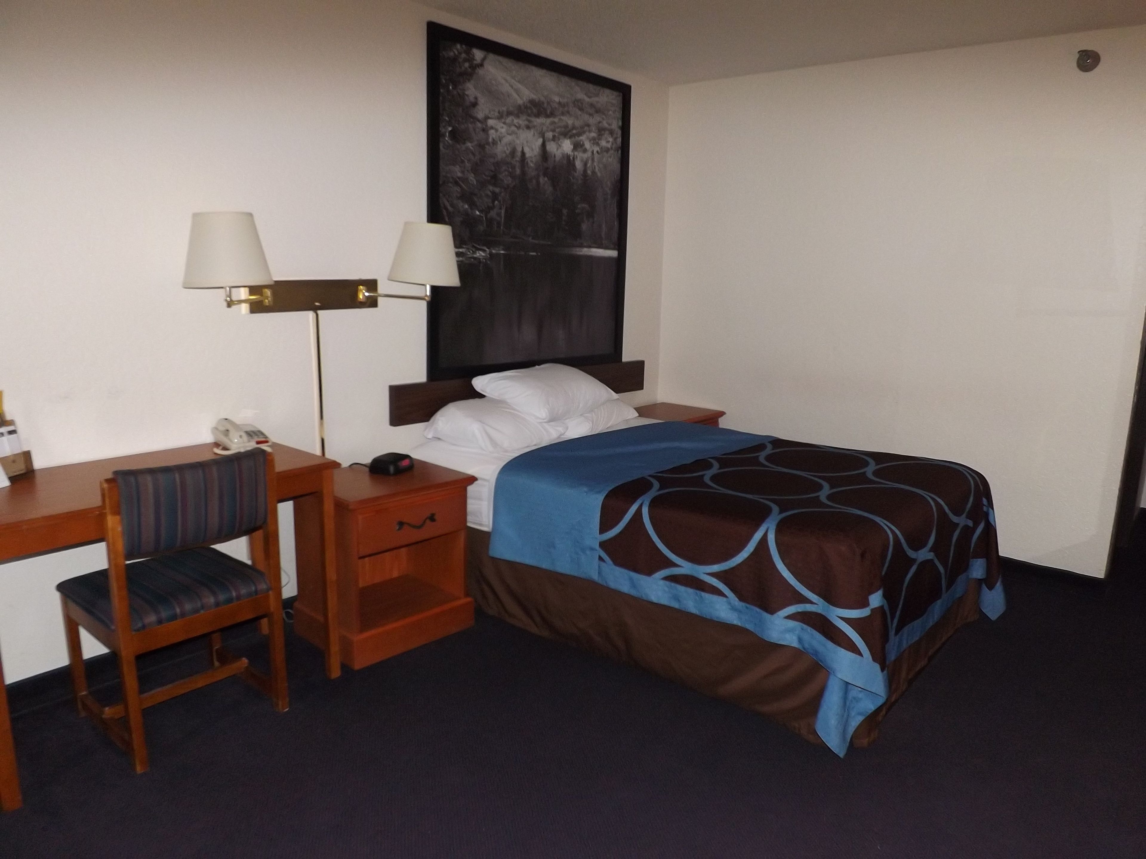 A hotel room with a bed, a desk, a chair, a nightstand, a lamp, and a painting on the wall.