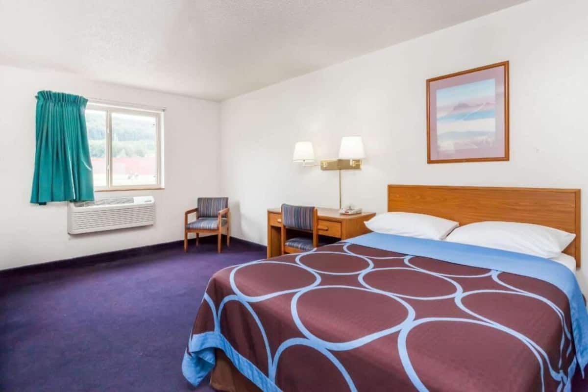 A hotel room with a bed, a desk, a chair, a window with curtains, an air conditioning unit, and a wall-mounted lamp.