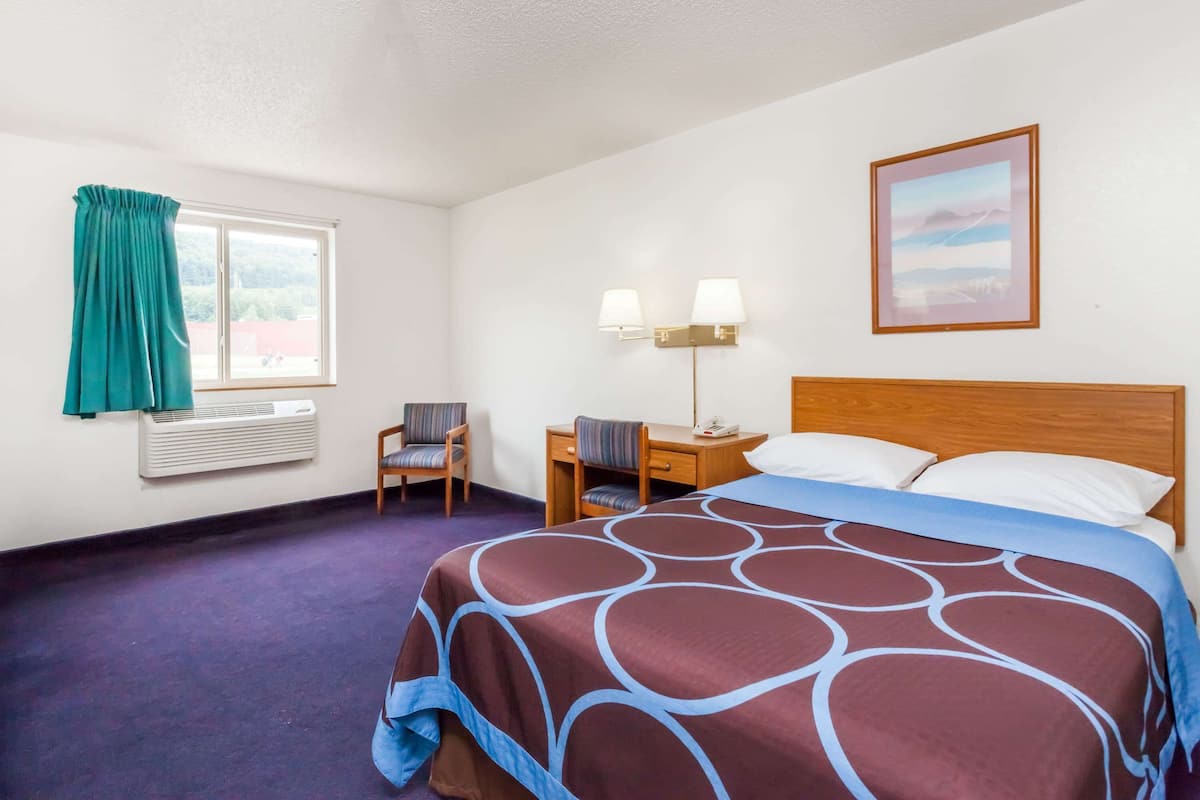 A hotel room with a large bed, a desk with a chair, a window with curtains, a wall-mounted lamp, and a painting.