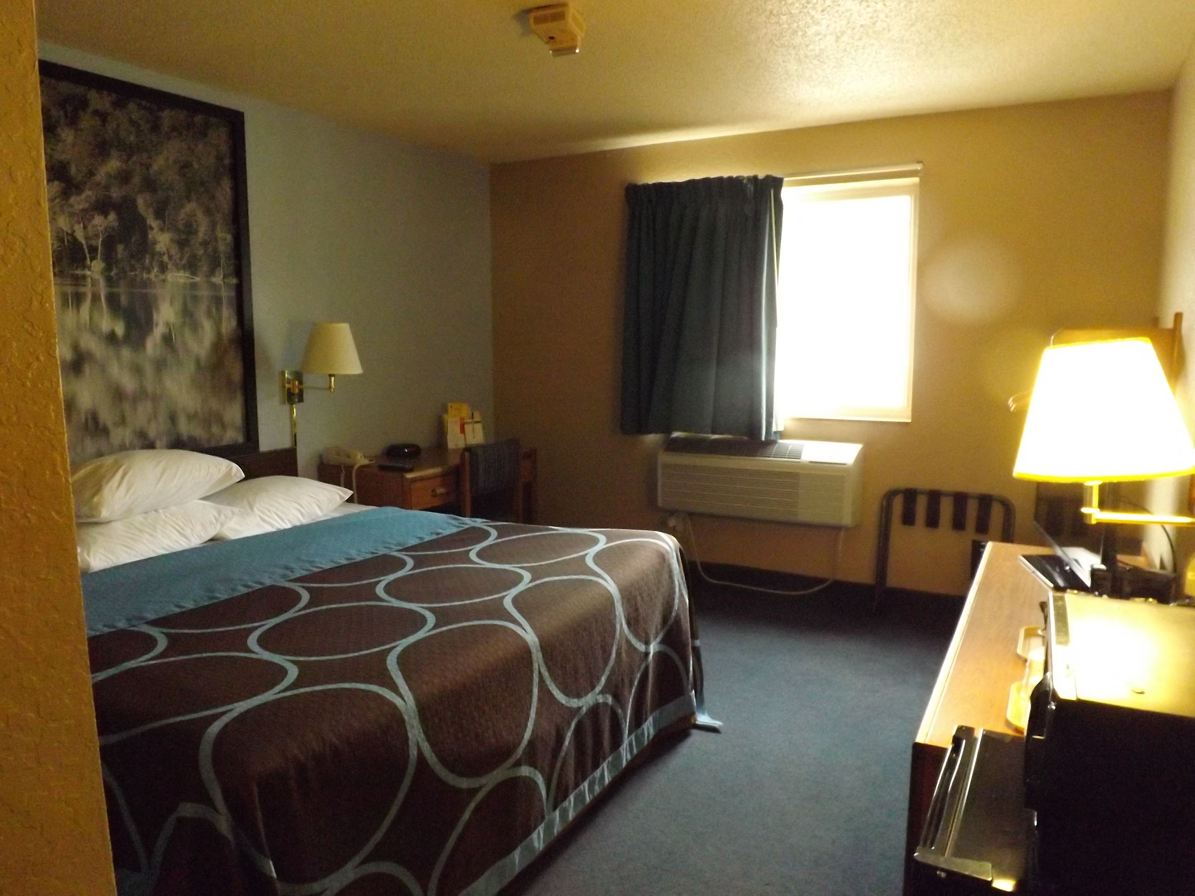 Habitación de hotel con cama, escritorio con lámpara, aire acondicionado y ventana con cortinas.