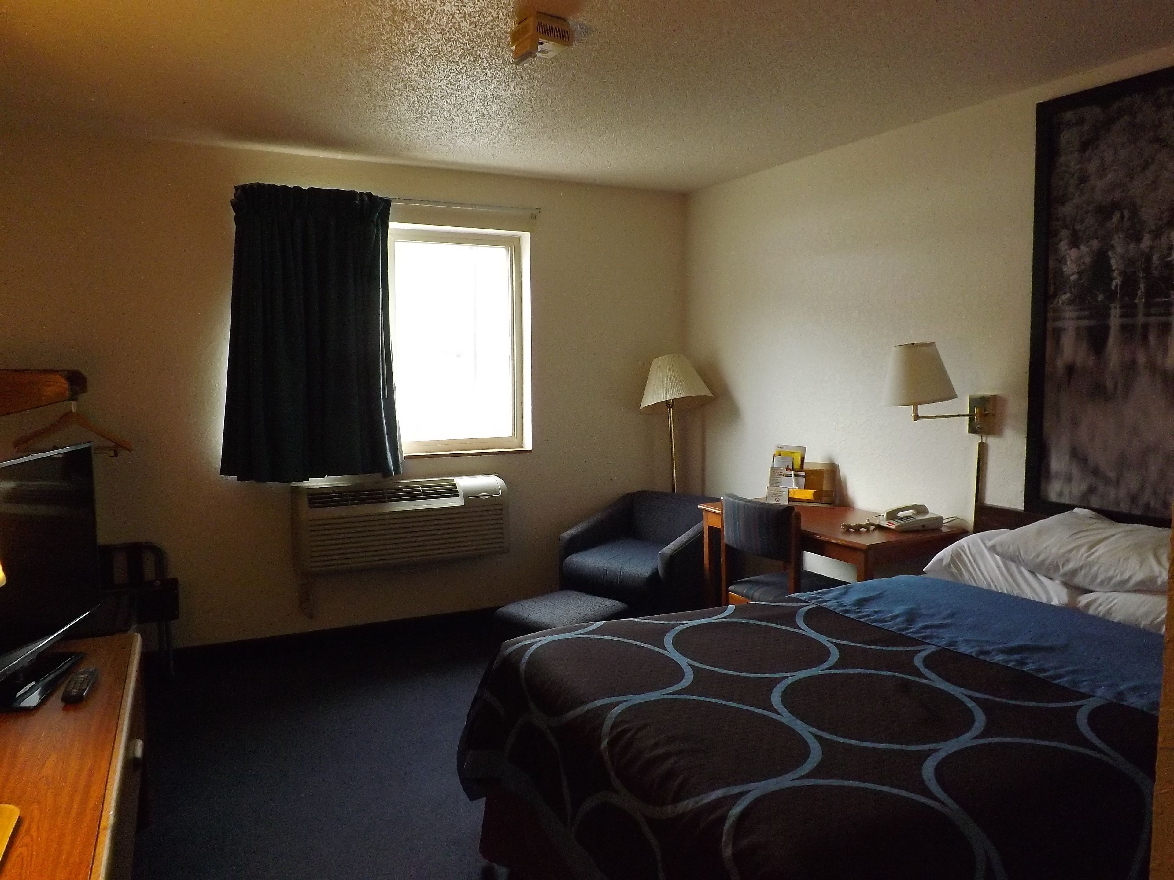 Ett hotellrum med en säng, ett skrivbord, en stol, ett bord med en lampa och ett fönster med gardiner.