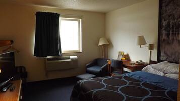 Ett hotellrum med en säng, ett skrivbord, en stol, ett bord med en lampa och ett fönster med gardiner.