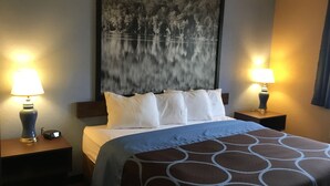 Quarto de hotel com cama, duas luminárias de cabeceira, um quadro abstrato grande na parede e uma mesa de cabeceira com relógio.