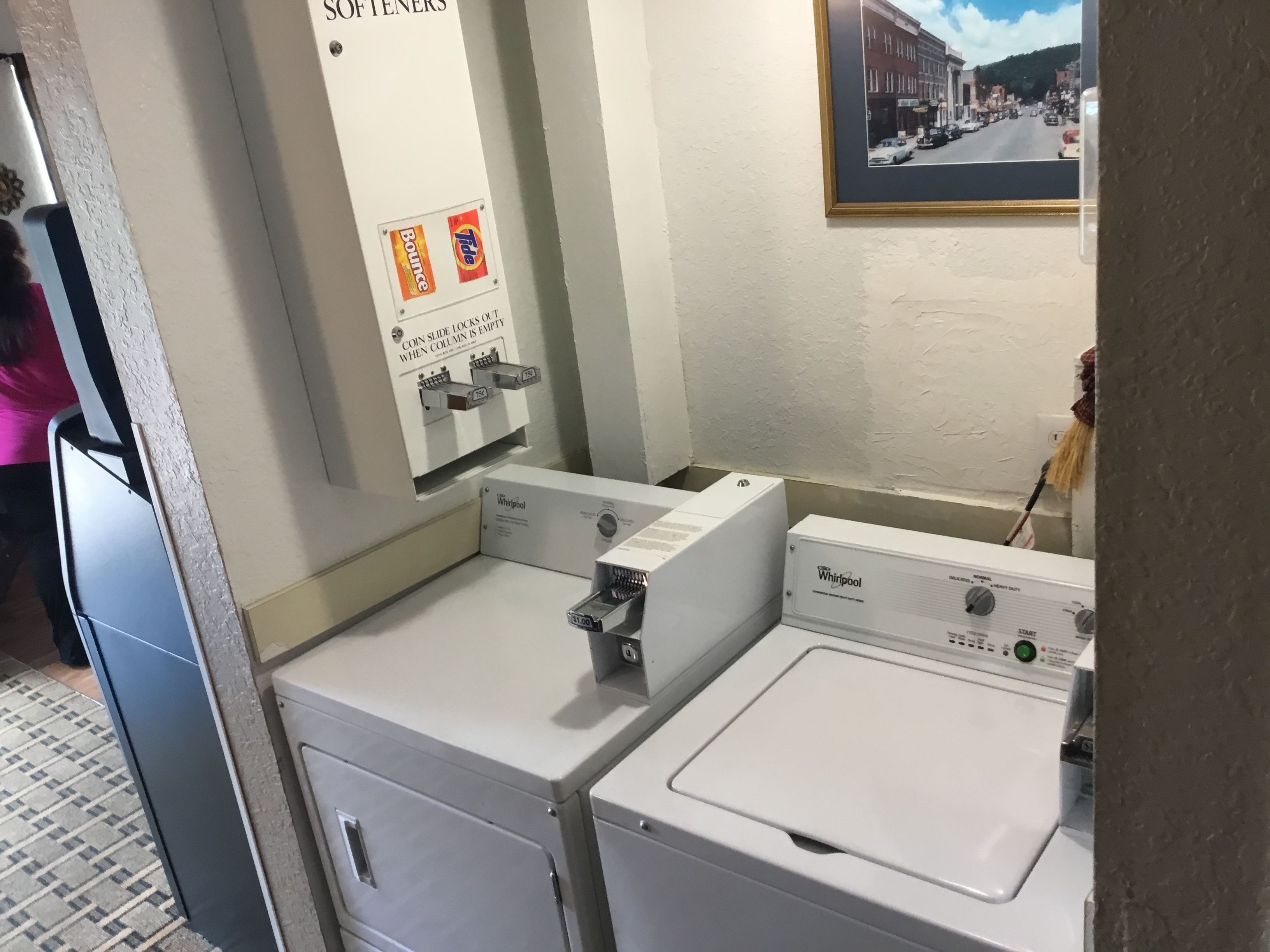 Et vaskerum med to Whirlpool vaskemaskiner, en møntåbning til vaskemiddel og et skilt, der angiver at blødgøringsmiddel er tilgængeligt.