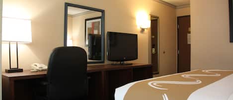 Habitación de hotel con cama, escritorio, silla, televisor y espejo.