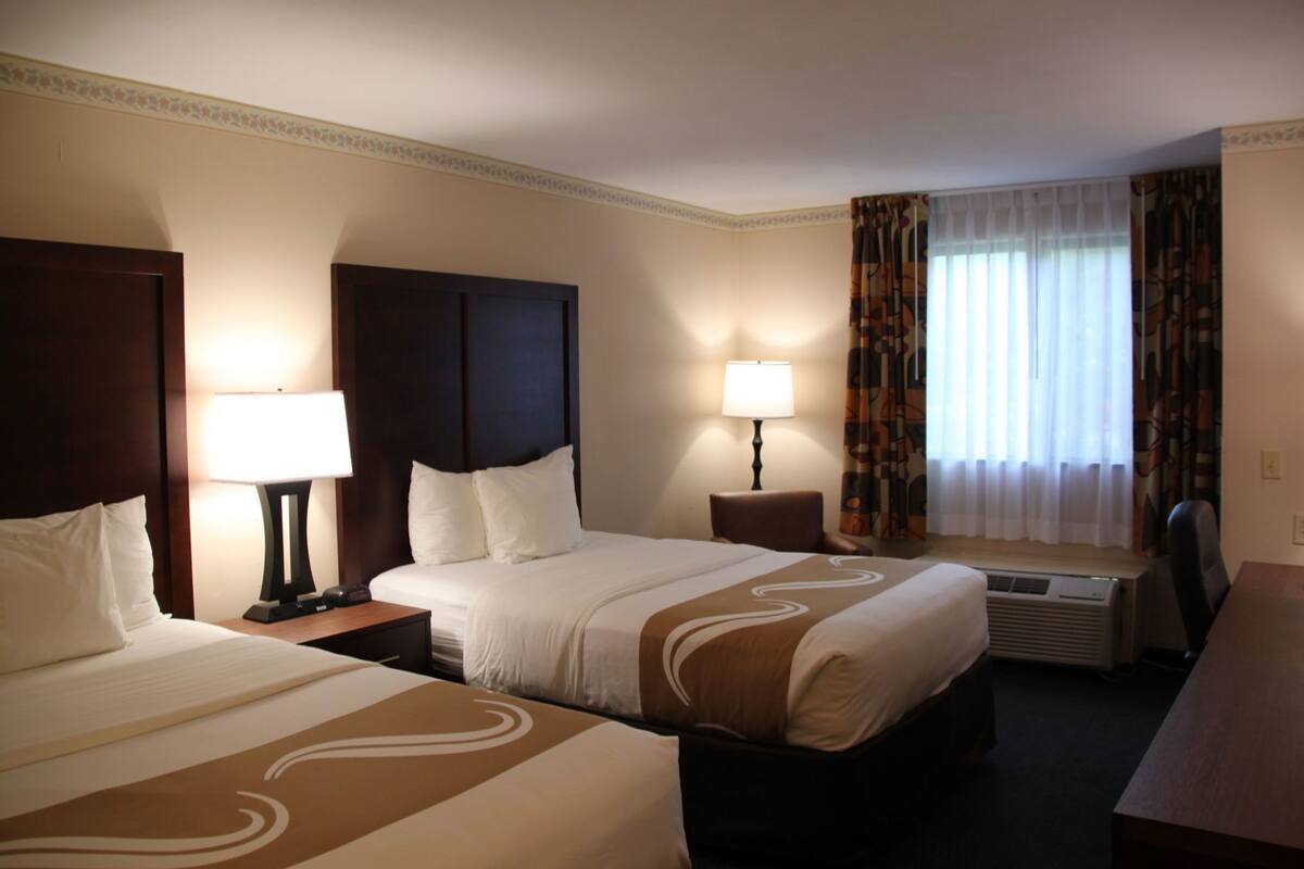 A hotel room with two beds, a desk, a chair, a window with curtains, and an air conditioning unit.