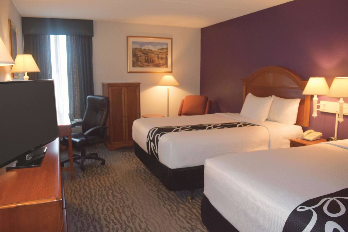 Habitación de hotel con dos camas, un escritorio con televisor, una silla, una lámpara y un cuadro en la pared.