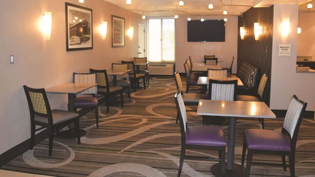 Un comedor de hotel con varias mesas y sillas, una alfombra estampada, luces fijadas a la pared y un letrero que indica los horarios del desayuno.