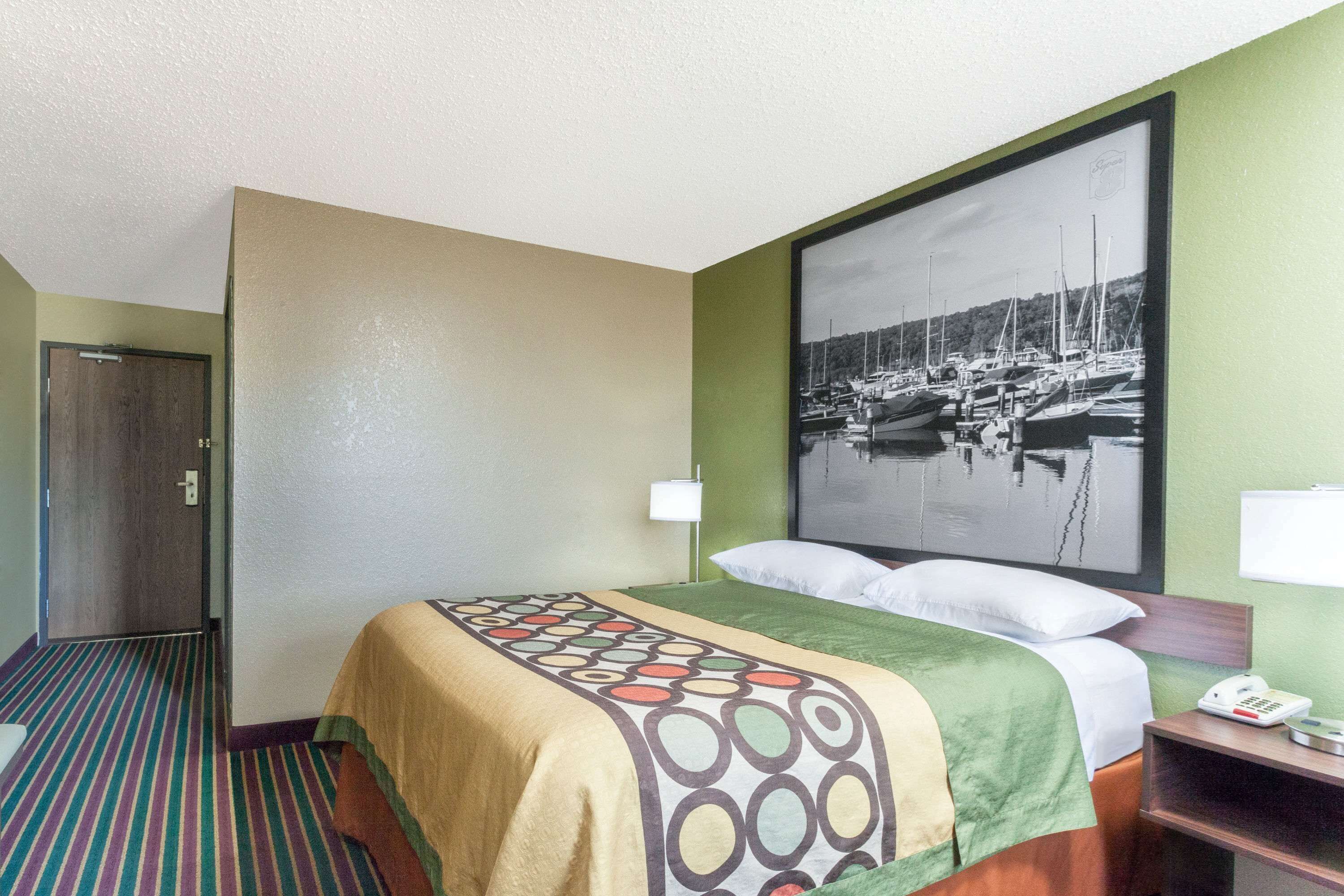 Quarto de hotel com cama, mesa de cabeceira, telefone e um quadro na parede retratando um porto.