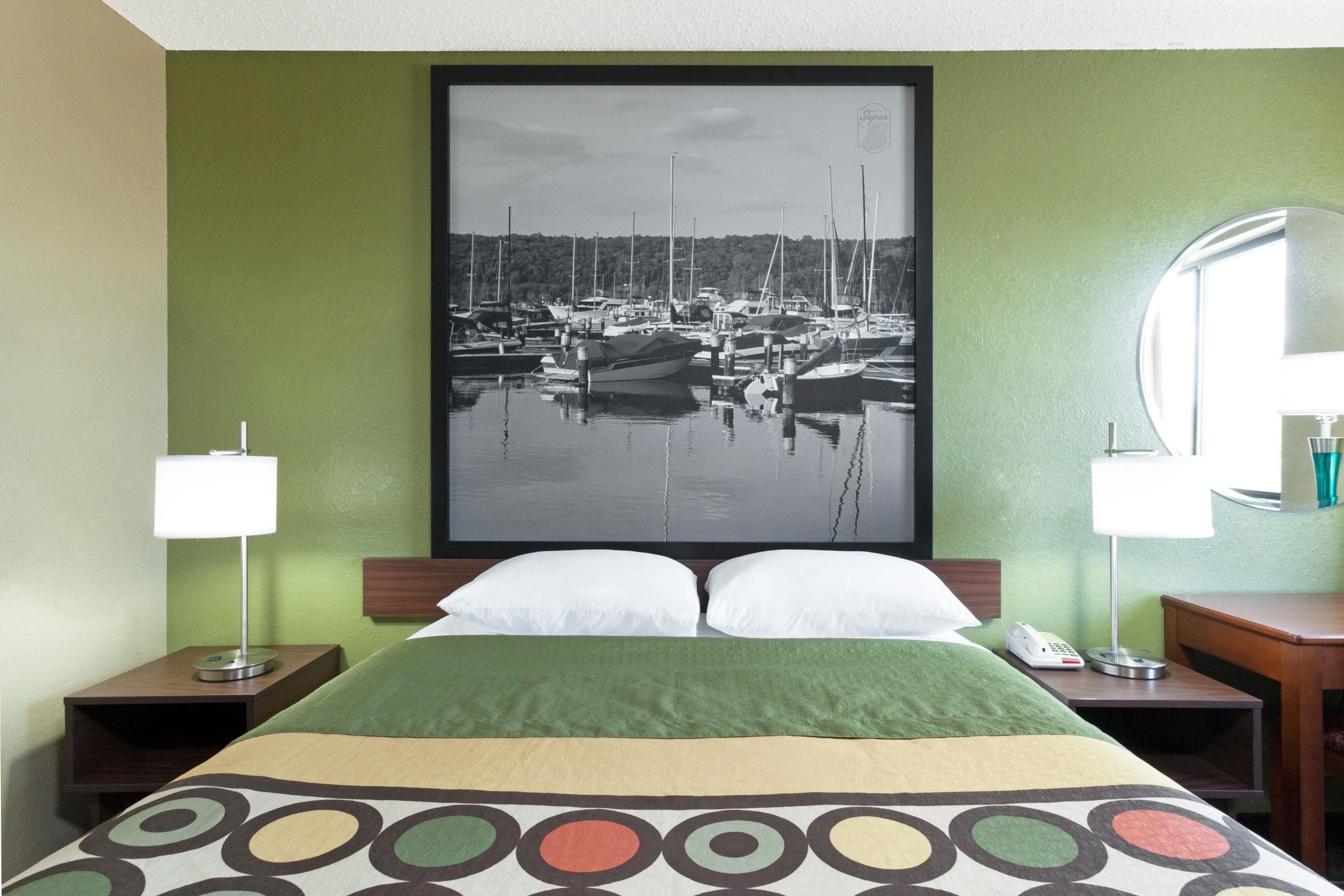 A hotel room with a bed, green walls, a framed photograph of a marina, two bedside lamps, and a round mirror.
