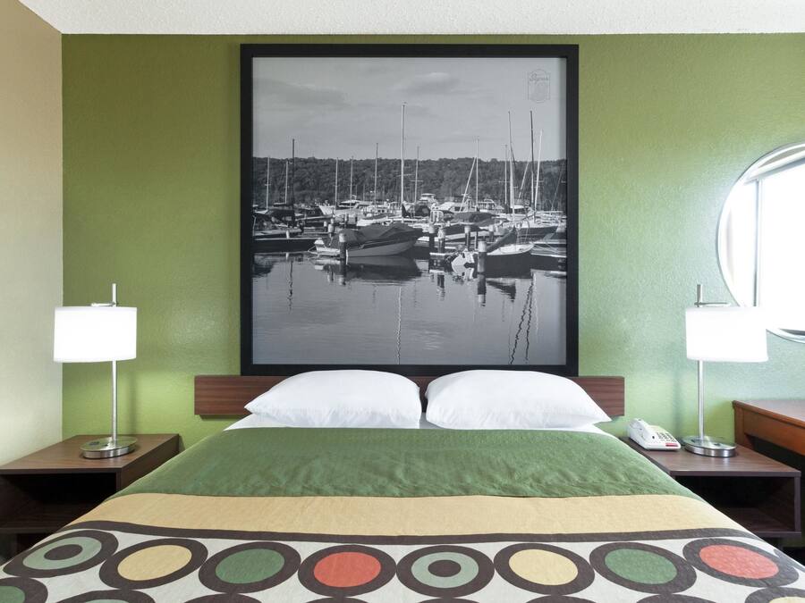 Ein Hotelzimmer mit Bett, grünen Wänden, einem gerahmten Foto eines Hafens, zwei Nachttischlampen und einem runden Spiegel.