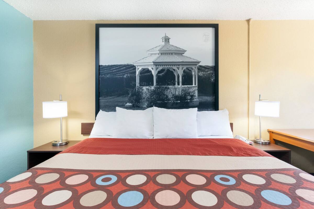Una habitación de hotel con una cama grande, dos mesitas de noche, una mesita auxiliar, un televisor montado en la pared y un cuadro con una imagen de un pabellón.