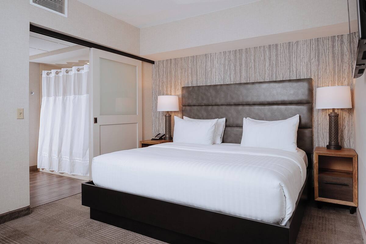 A hotel room with a large bed, two bedside tables with lamps, a wooden headboard, and a view of the bathroom.