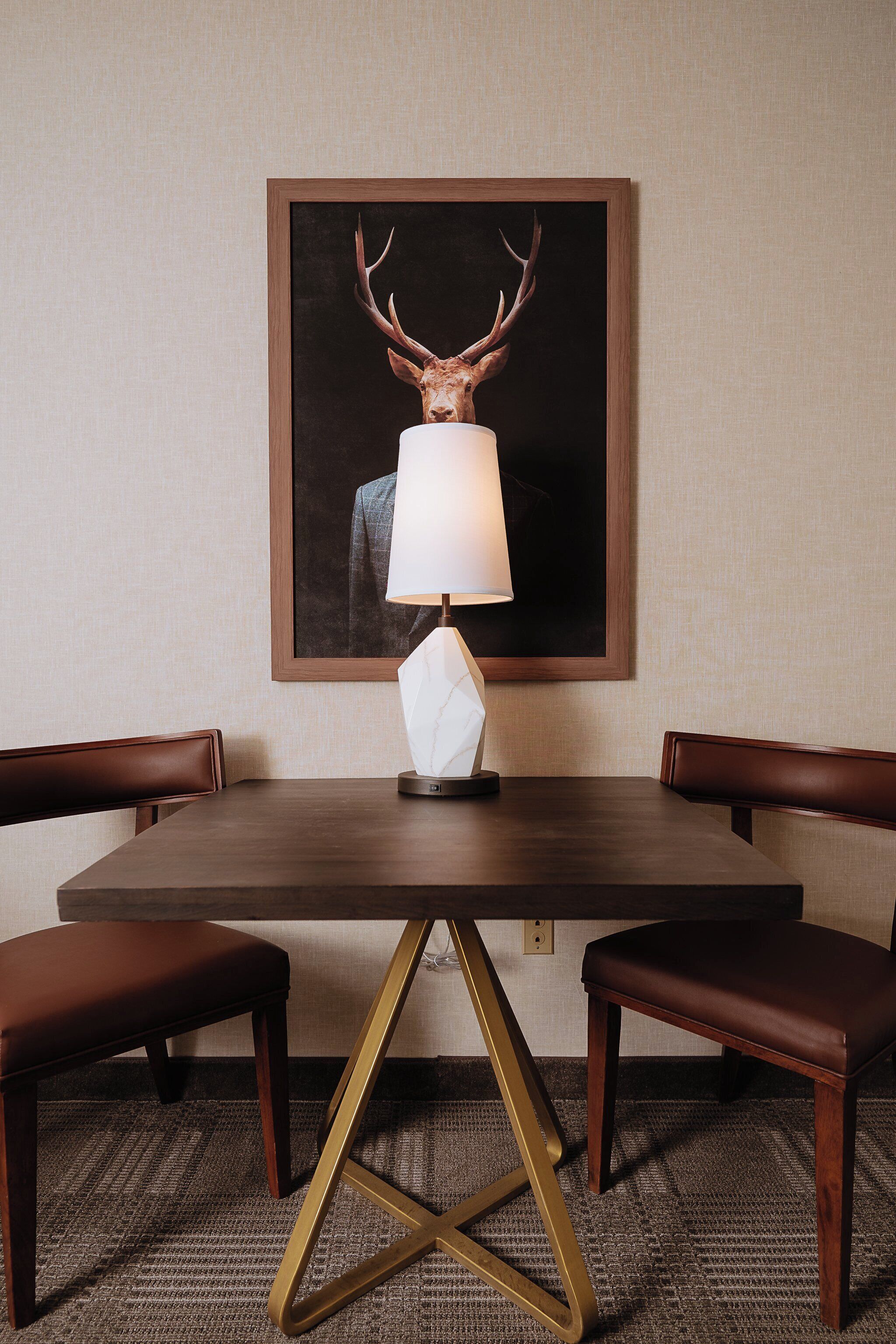 A dining table with a framed deer head artwork above it, a modern lamp, and leather chairs.