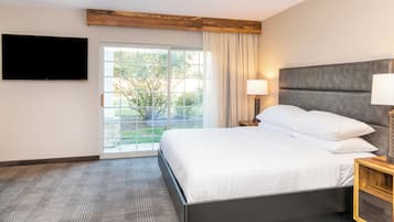Una habitación de hotel con una cama, un televisor montado en la pared, una ventana con cortinas y una mesita de noche con lámpara.