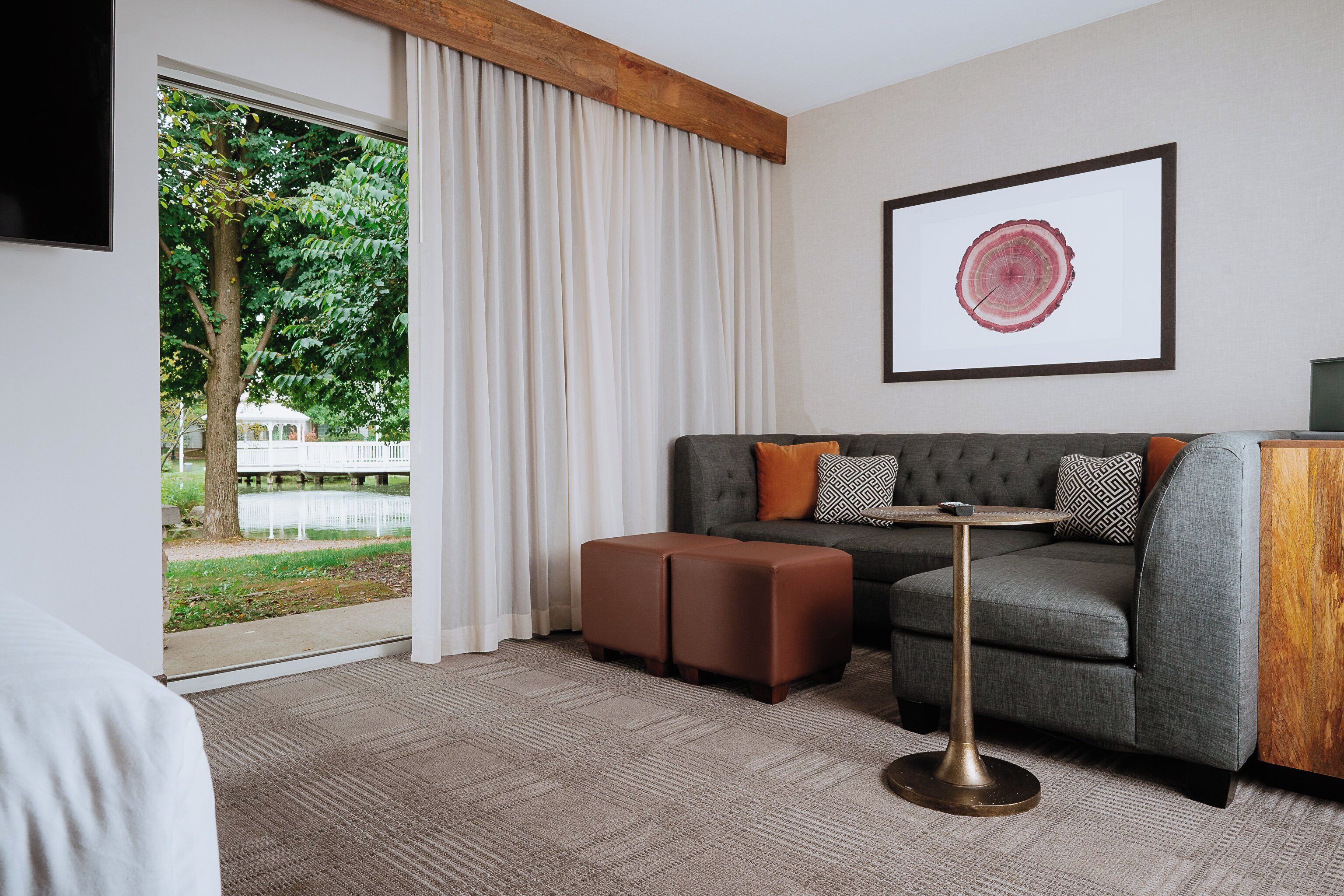 A modern hotel room with a grey sofa, brown ottoman, and a round table with a gold base. A large window with white curtains offers a view of a tree and a white building outside.