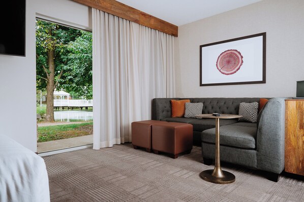 Una habitación de hotel moderna con un sofá gris, un reposapiés marrón y una mesa redonda de base dorada. Un amplio ventanal con cortinas blancas ofrece vistas a un árbol y un edificio blanco en el exterior.