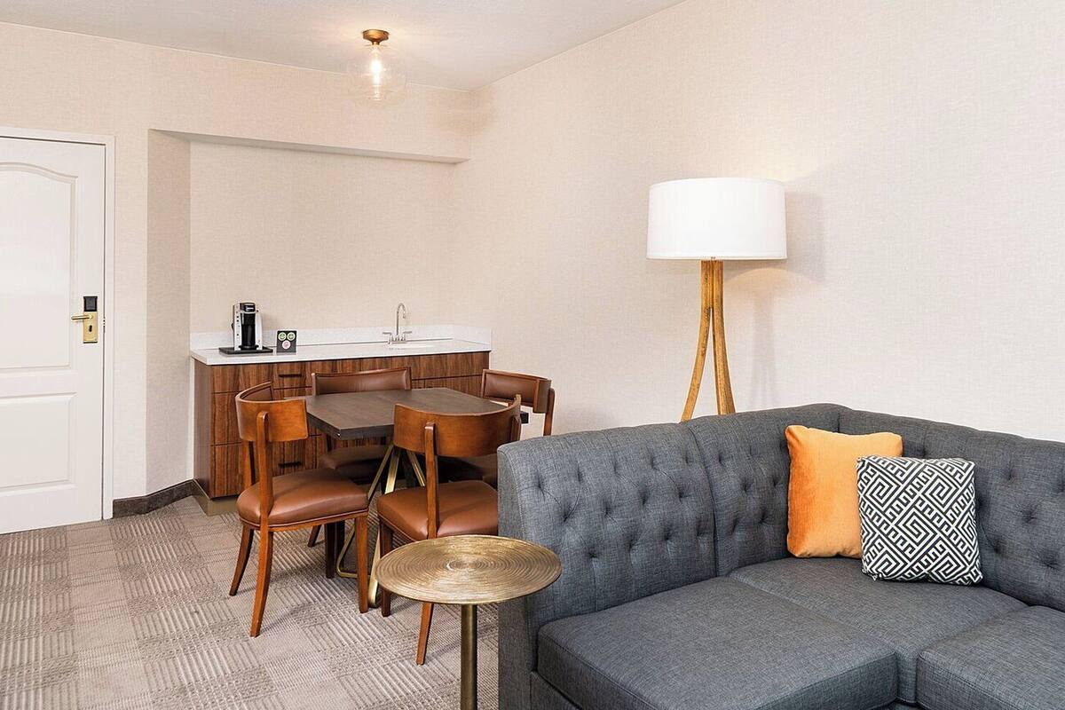 A hotel room with a grey sofa, a small round table, wooden chairs, a white door, a kitchenette with a sink, and a lamp.