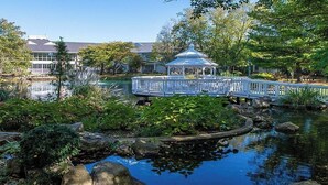 Ein Pavillon mit weißem Dach und Geländer, umgeben von einem Teich und üppiger Bepflanzung.