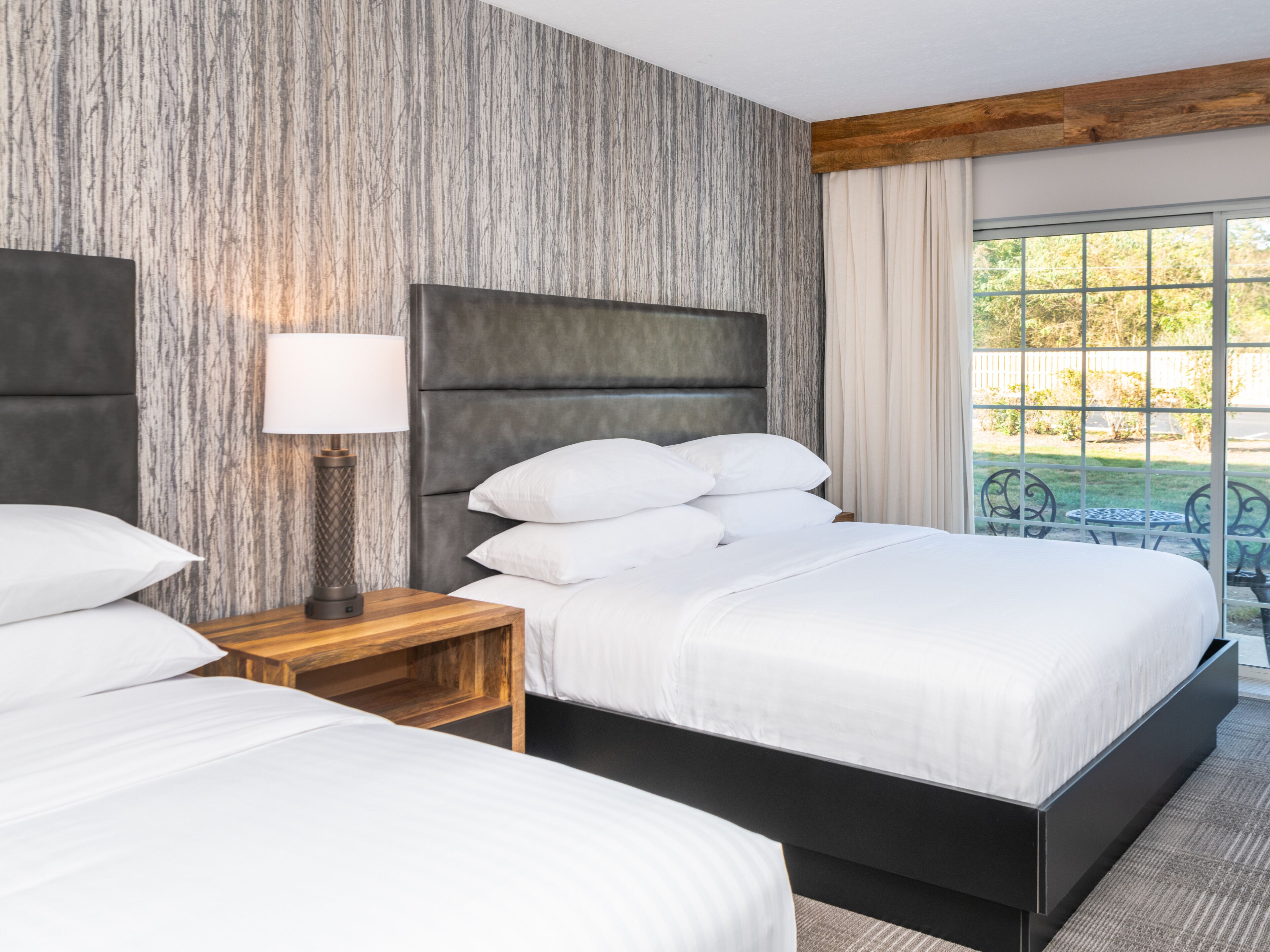 A hotel room with a large bed, a bedside table with a lamp, and a view of the outdoors through a window.