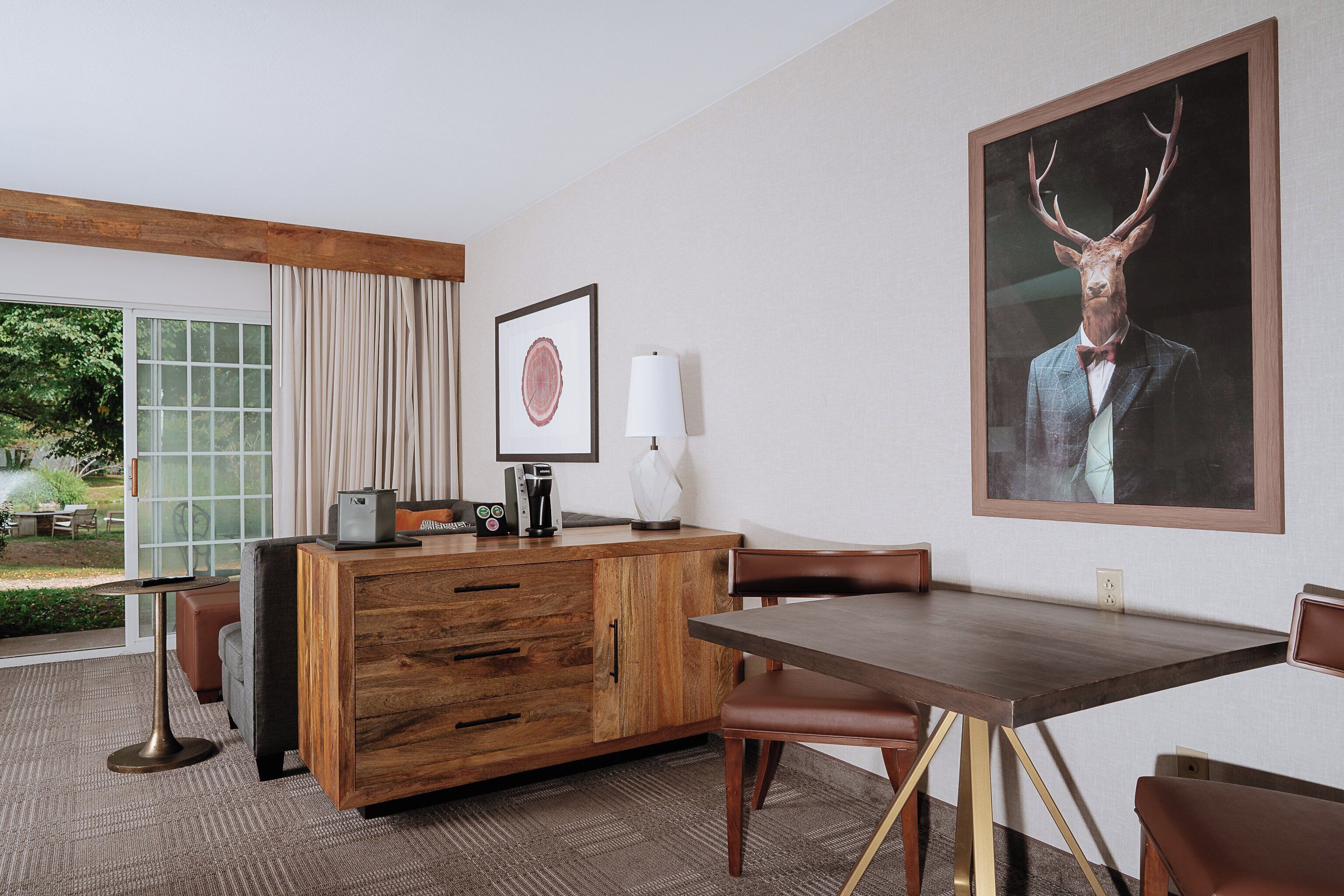 A hotel room with a wooden desk, leather chairs, a large framed deer head artwork, and a view of greenery outside.
