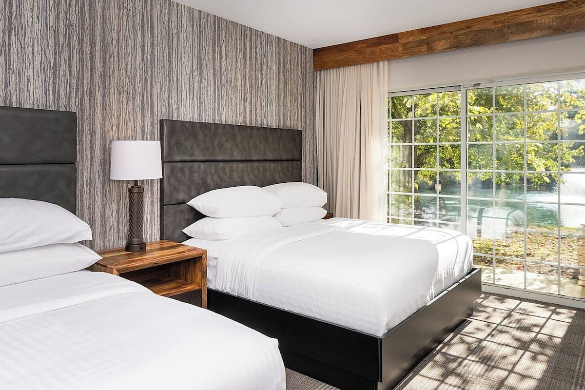 A hotel room with a large bed, a bedside table, a lamp, and a view of greenery outside.