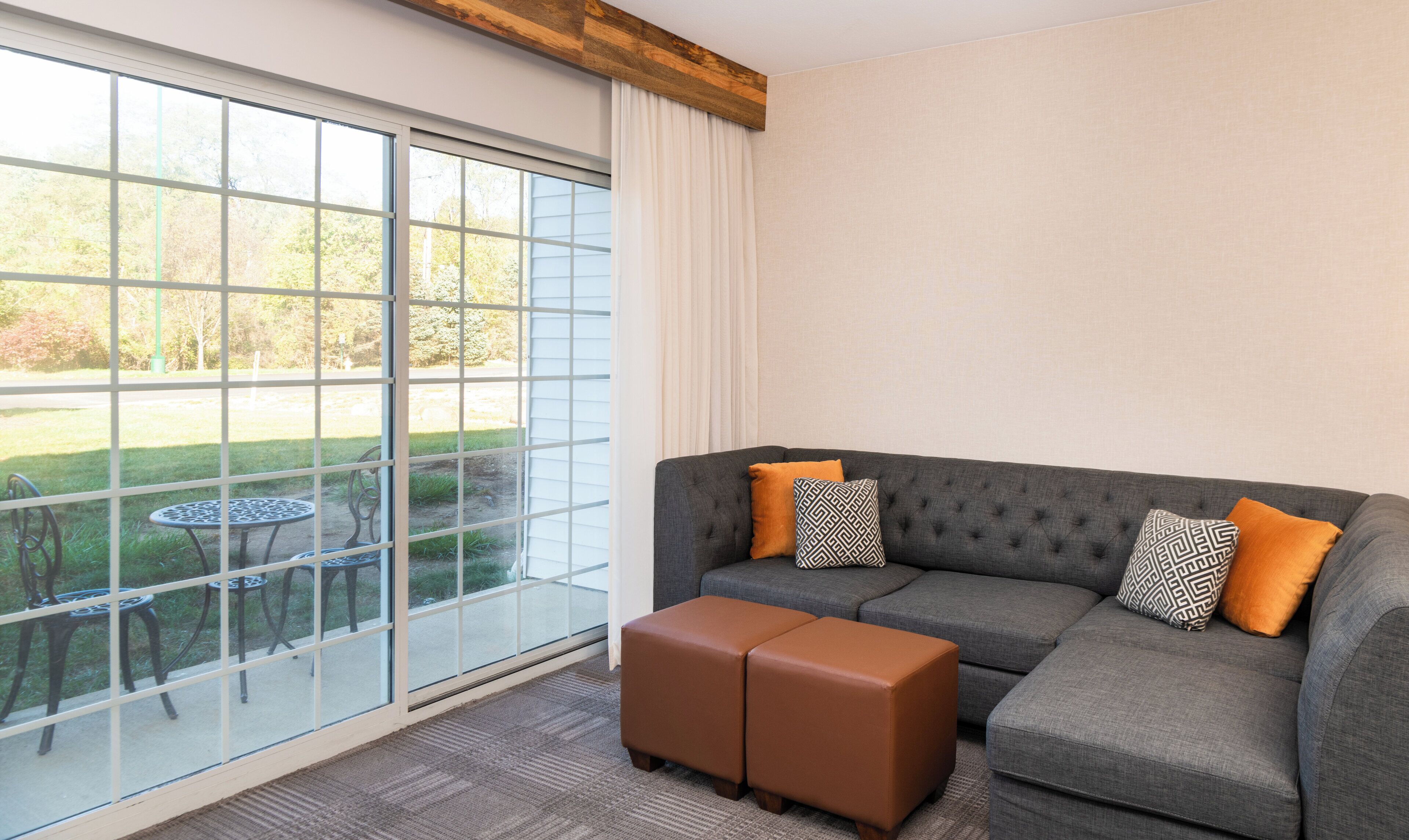 A modern living room with a grey sectional sofa, two ottomans, and a small round table. A large window offers a view of a garden and a street.