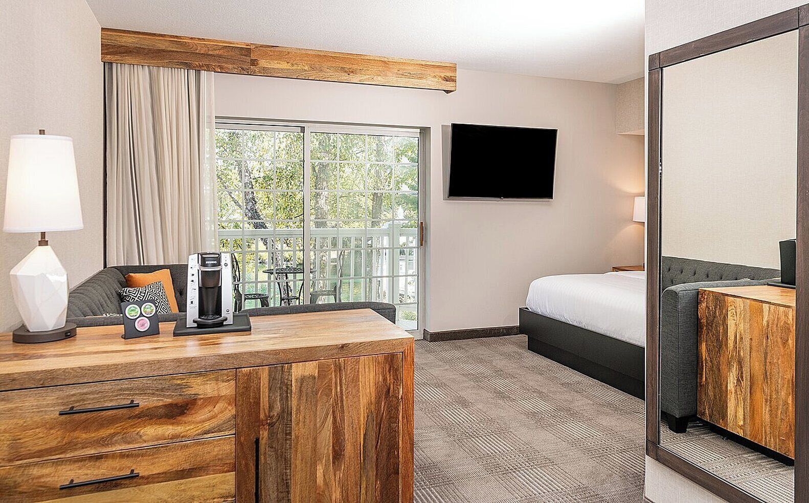 A hotel room with a wooden desk, a bed, a TV, and a large window with a view of greenery.