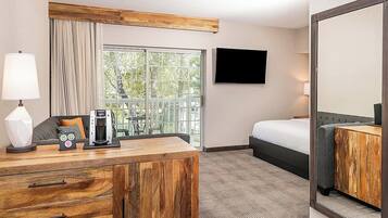 A hotel room with a wooden desk, a bed, a TV, and a large window with a view of greenery.