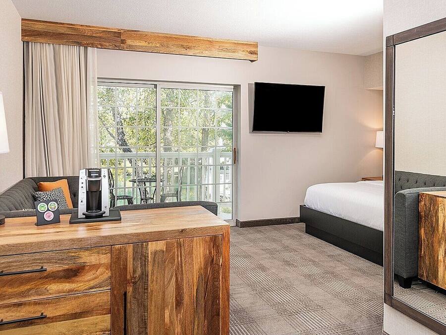 A hotel room with a wooden desk, a bed, a TV, and a large window with a view of greenery.