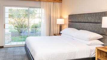 A modern hotel room with a large bed, a wooden headboard, a nightstand with a lamp, and a view of greenery through a sliding glass door.