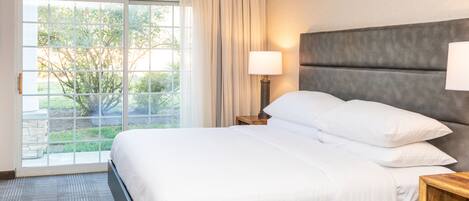Una habitación de hotel moderna con una cama grande, un cabecero de madera, una mesita de noche con lámpara y vistas a zonas verdes a través de un ventanal corredero.