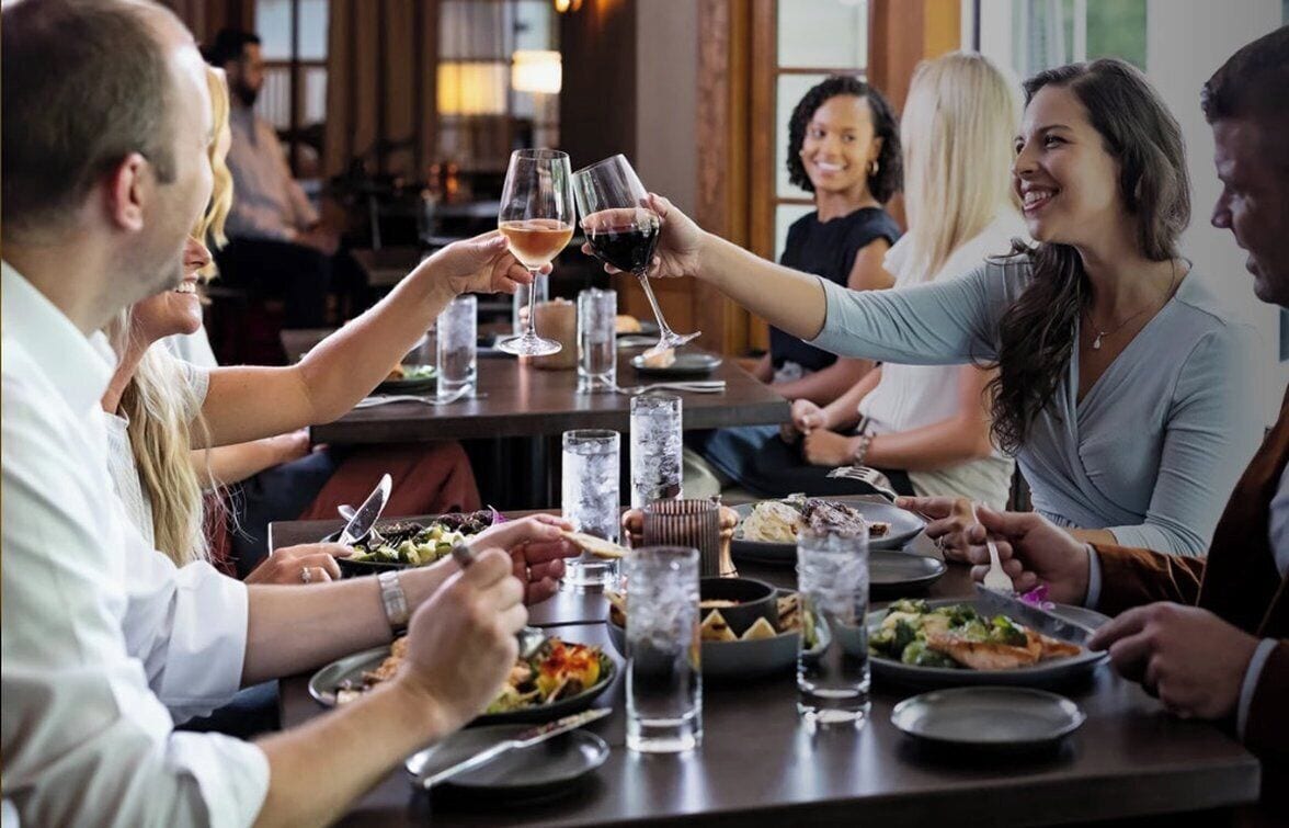 A group of people dining at a restaurant with multiple tables, each with plates of food and glasses of beverages.
