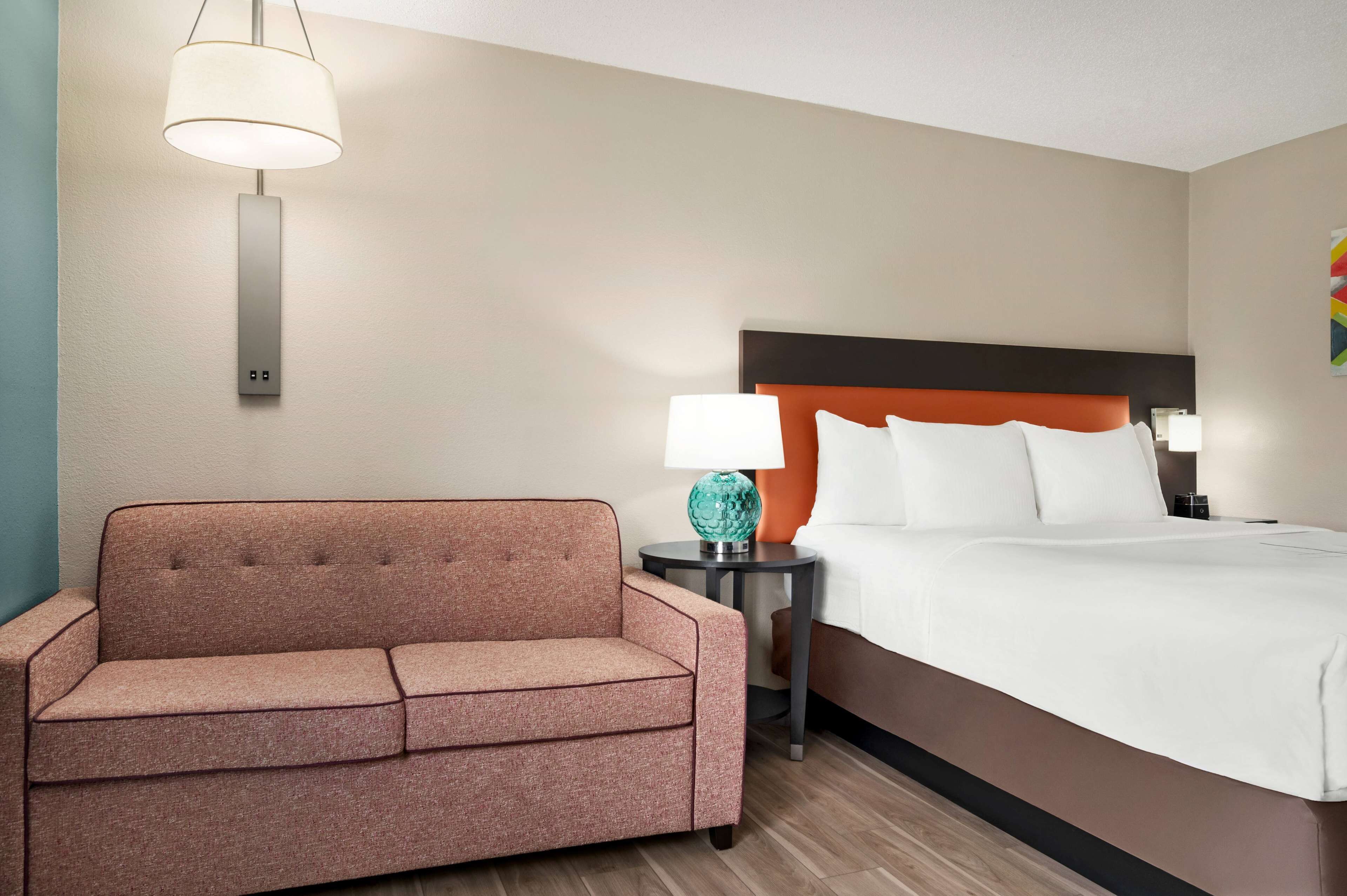 A hotel room with a bed, a sofa, a nightstand with a lamp, and a wall-mounted light fixture.