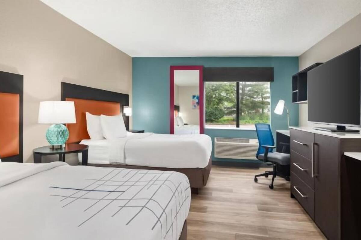 A hotel room with a large bed, a smaller bed, a desk with a TV, a chair, and a view of the outdoors.