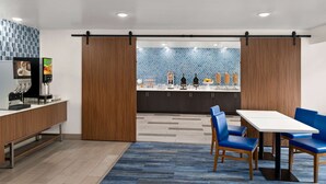 Een moderne keuken met een koffieapparaat, een koelkast en een toonbank met een koffiepresentatie. Er is een eethoek met een tafel en blauwe stoelen.