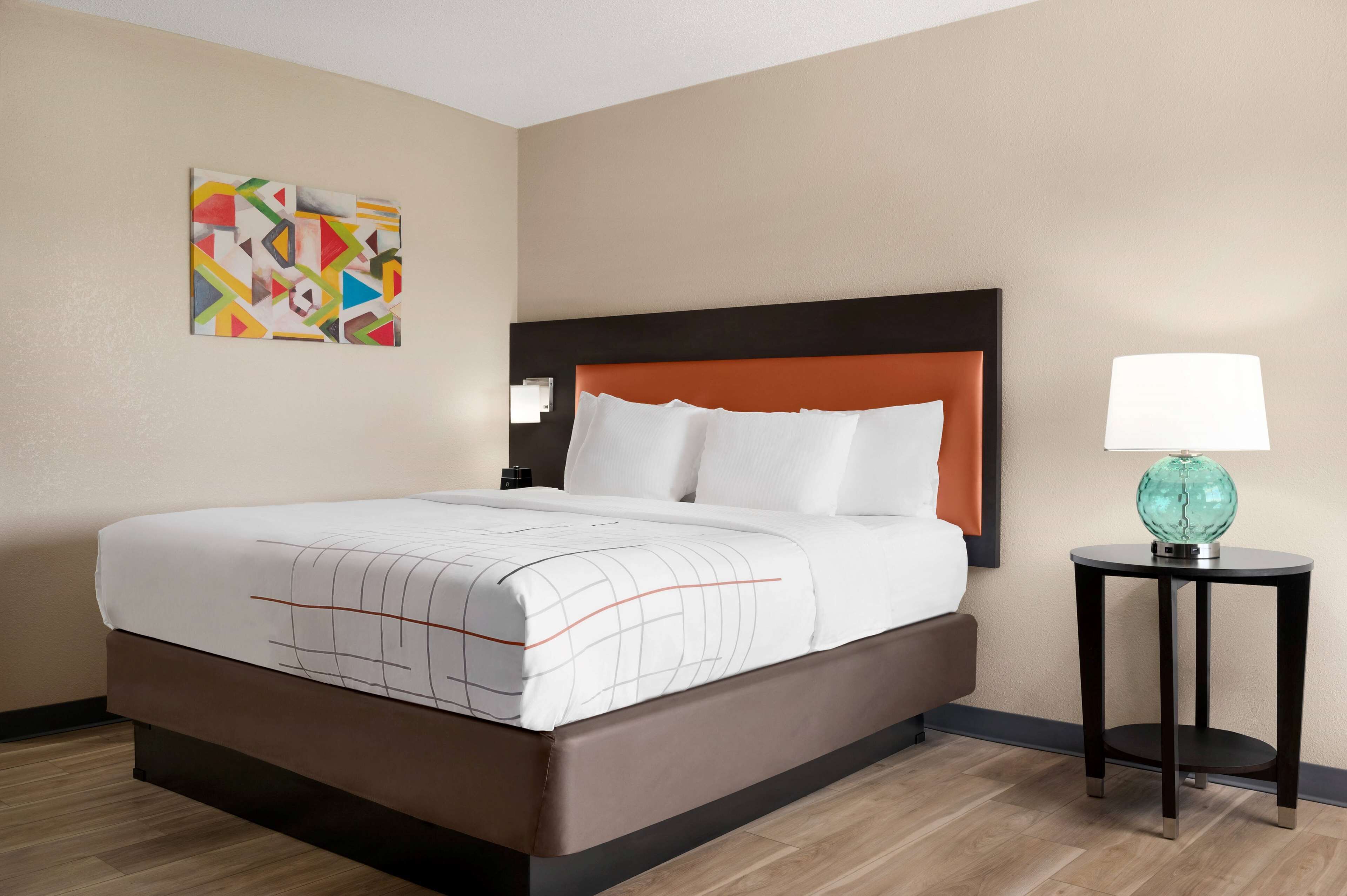 A hotel room with a bed, a nightstand, a lamp, and a wall art piece.