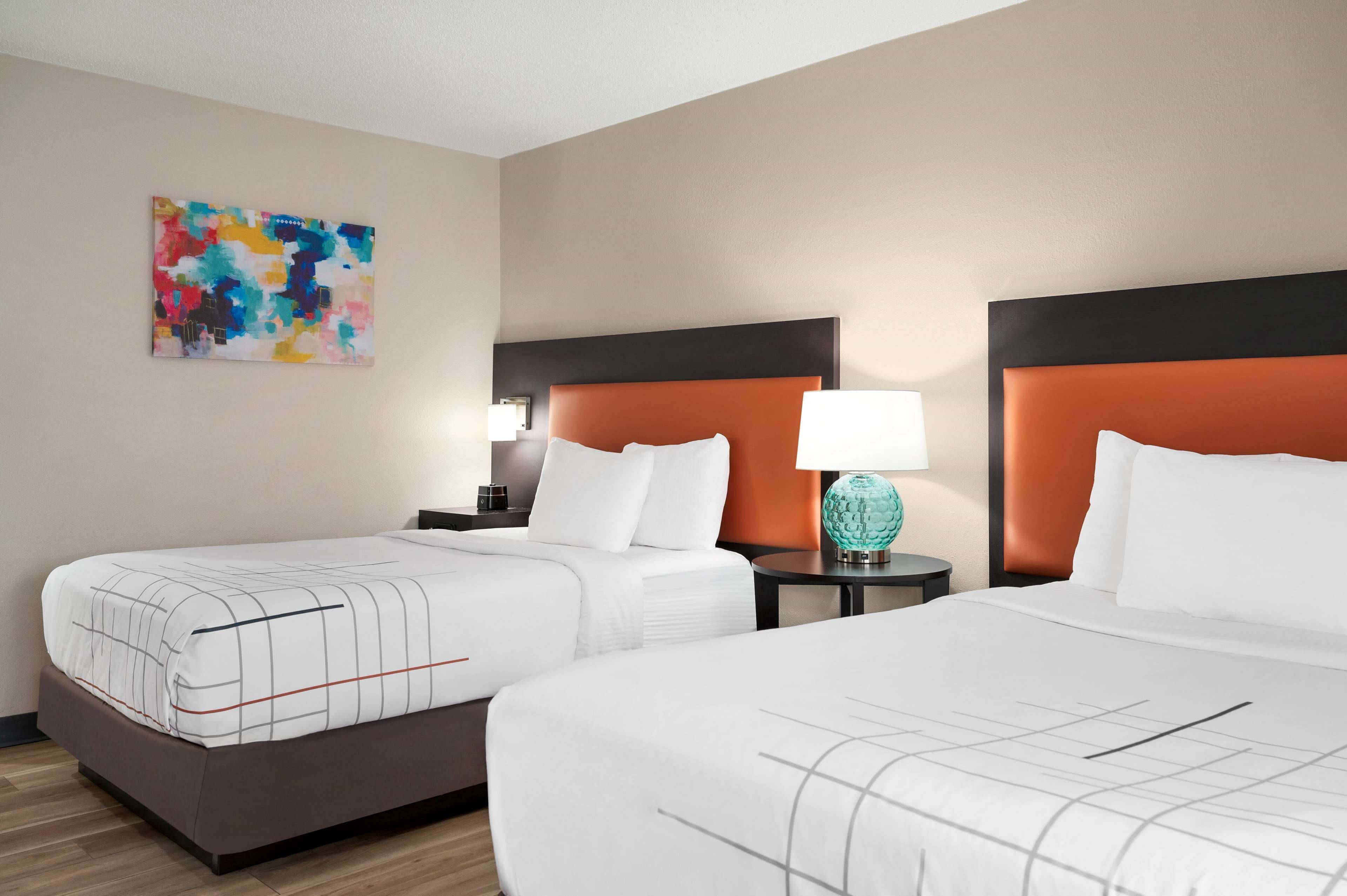 A hotel room with two beds, each with a headboard, a bedside table with a lamp, and a colorful abstract painting on the wall.