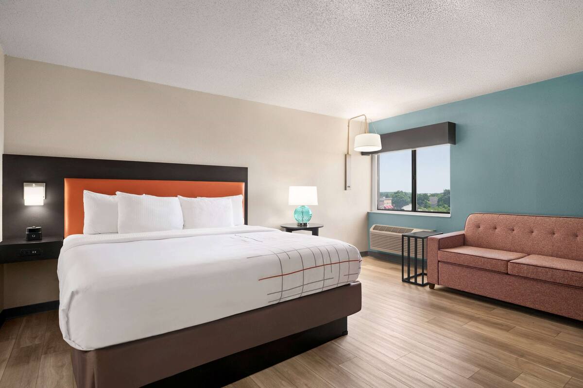A hotel room with a large bed, a sofa, a nightstand with a lamp, and a window with a view.