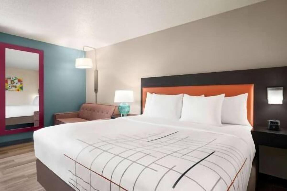 A hotel room with a large bed, a nightstand, a chair, a mirror, and a wall with a painting.